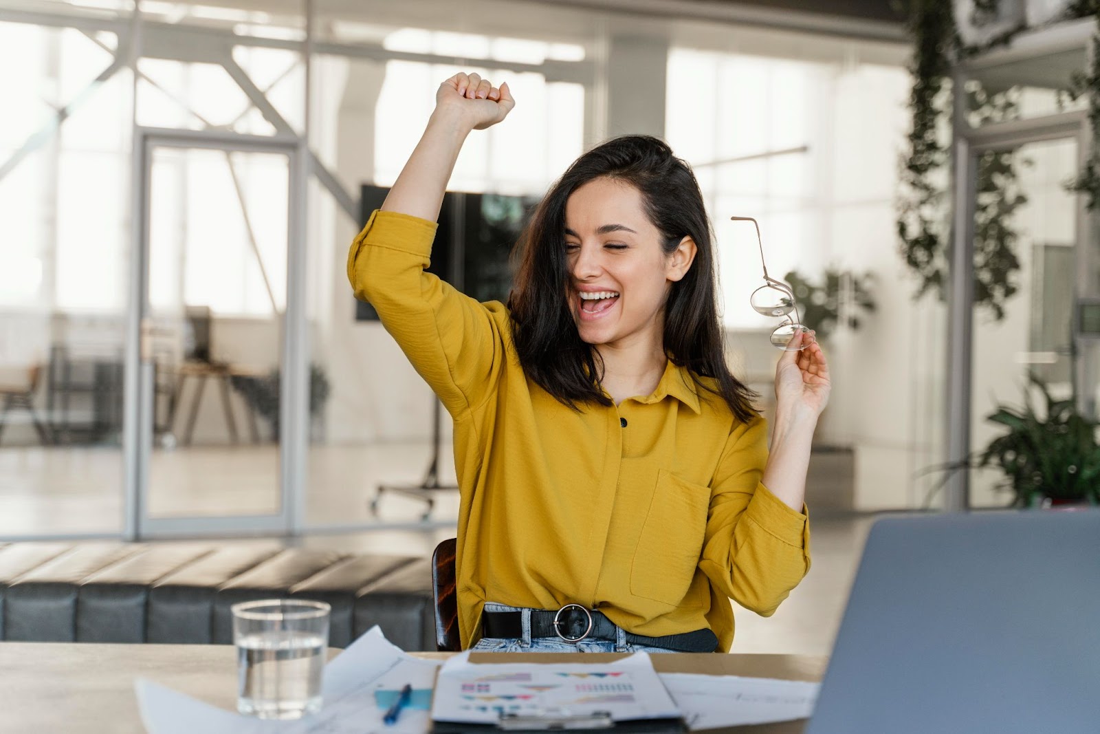 Mulher com camisa amarela sentada em uma cadeira enquanto está trabalhando, comemora e sorri, expressando felicidade pelo recebimento do decimo terceiro 2023. Em sua mão segura um óculos de grau.