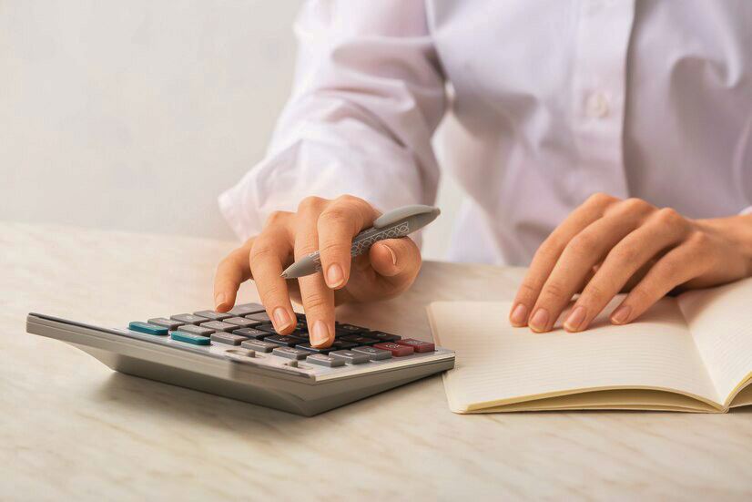 Mão de uma mulher apertando botões de uma calculadora enquanto segura uma caneta para calcular o decimo terceiro 2023. Sua outra mão está em cima de um caderno de anotações.