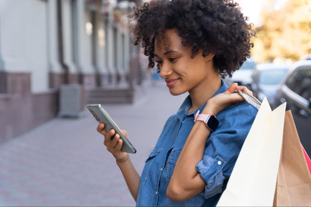 Mulher sorrindo e andando na rua com camisa azul. Ela segura sacolas de compras enquanto mexe em seu celular.