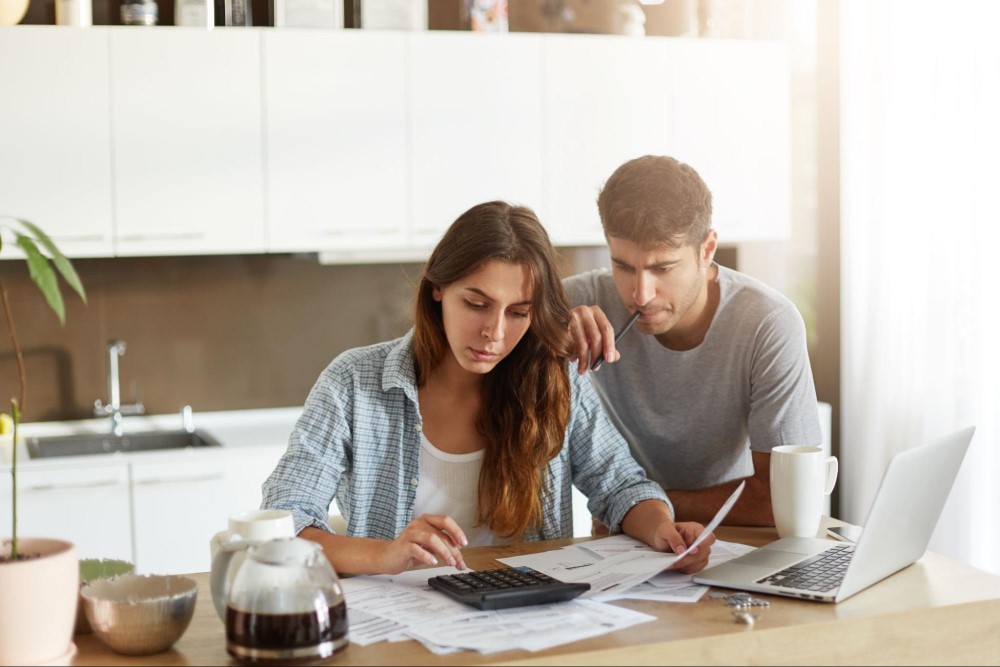 Casal sentado em volta da mesa da cozinha. Enquanto o homem morde um lápis pensativo, a mulher usa uma calculadora para fazer as contas do payback. Ao redor, vemos café e um notebook.