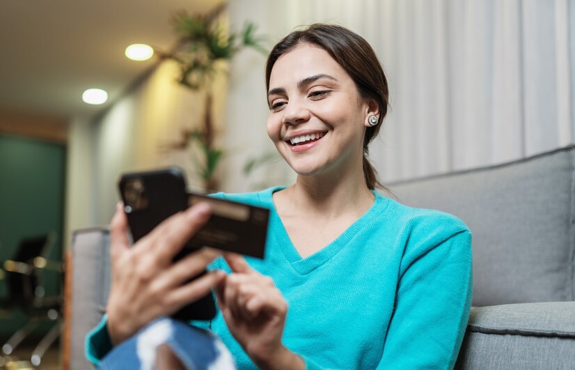 Mulher com blusa azul, sentada em um sofá, sorri enquanto segura seu celular e um cartão de crédito.