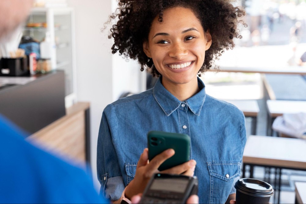 Mulher negra com camisa jeans, sorrindo em um estabelecimento, enquanto paga por aproximação o seu café.