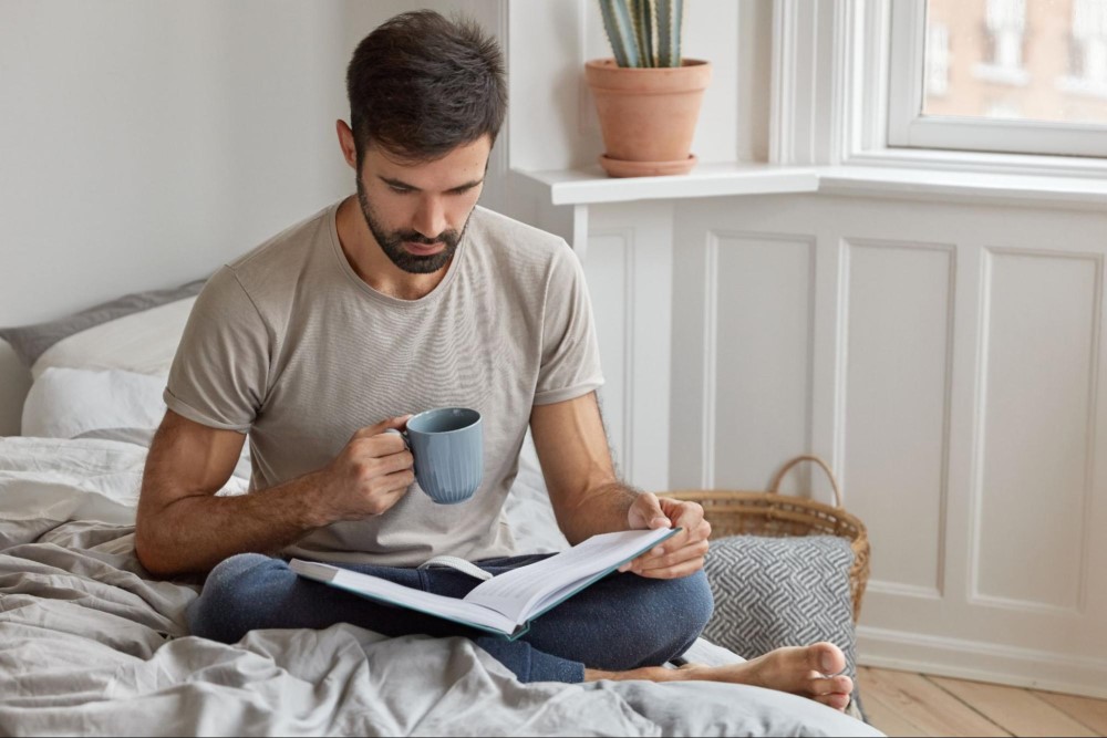 Homem branco sentado com as pernas cruzadas em uma cama, enquanto segura uma xícara de café e lê um livro. Ao redor, vemos uma parede branca, janela, vaso com cacto e almofadas.
