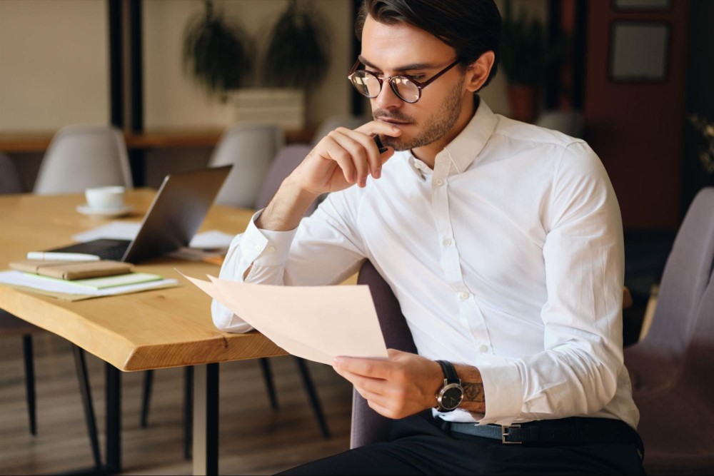 Homem com roupa social, com expressão de pensativo, sentado segurando um papel. Em sua volta vemos uma mesa, notebook, xícara de café.