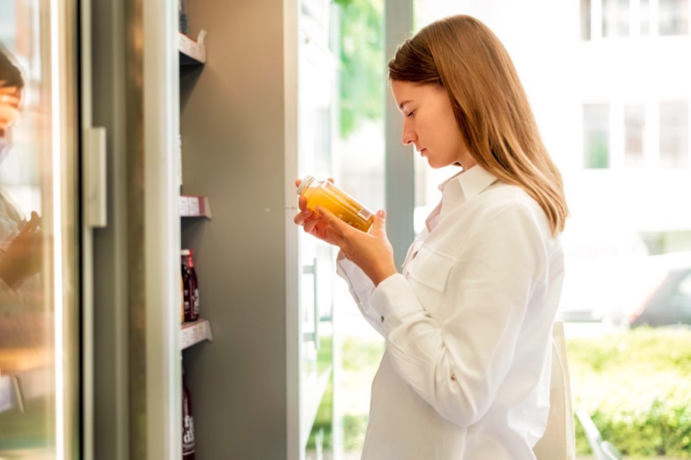 Mulher branca vestindo uma blusa branca está segurando uma garrafa, que contém um líquido amarelo, enquanto lê o rótulo. À sua frente estão prateleiras com sucos de variados sabores.