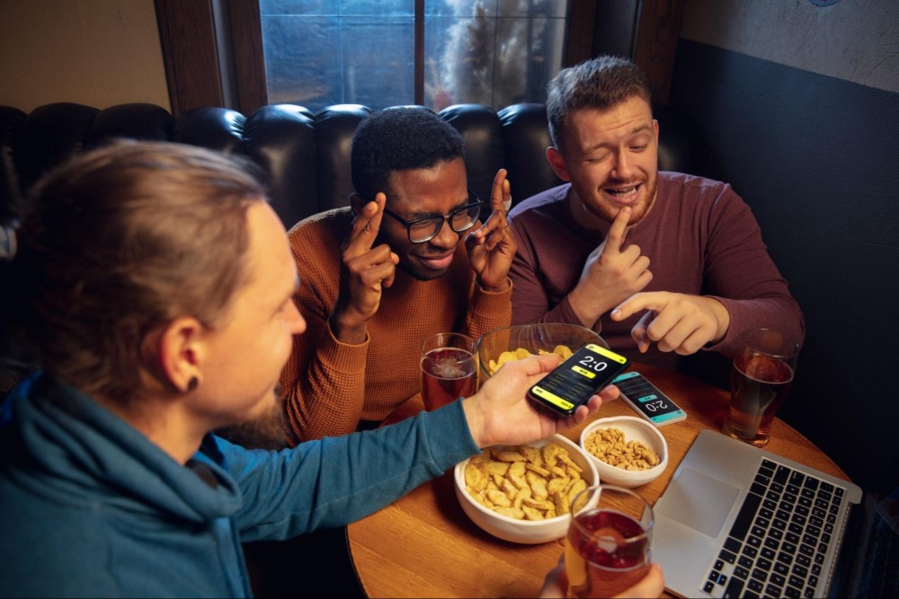 Três homens estão sentados em um sofá assistindo uma partida de esporte. Um deles está segurando seu celular e mostrando para o outro o resultado, enquanto o terceiro está cruzando os dedos em sinal de sorte. Na mesa vemos aperitivos, copos com bebida e um notebook.