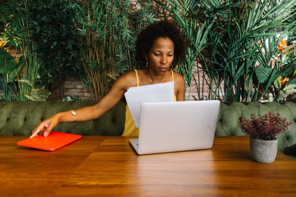 Uma mulher negra sentada em um banco almofadado, em frente a uma mesa de madeira. Sobre a mesa, estão um laptop e um vaso de planta. A mulher segura algumas folhas de papel na mão esquerda, enquanto coloca um tablet com capa vermelha sobre a mesa. Atrás dela, há várias plantas cobrindo uma parede de tijolinhos de granito.