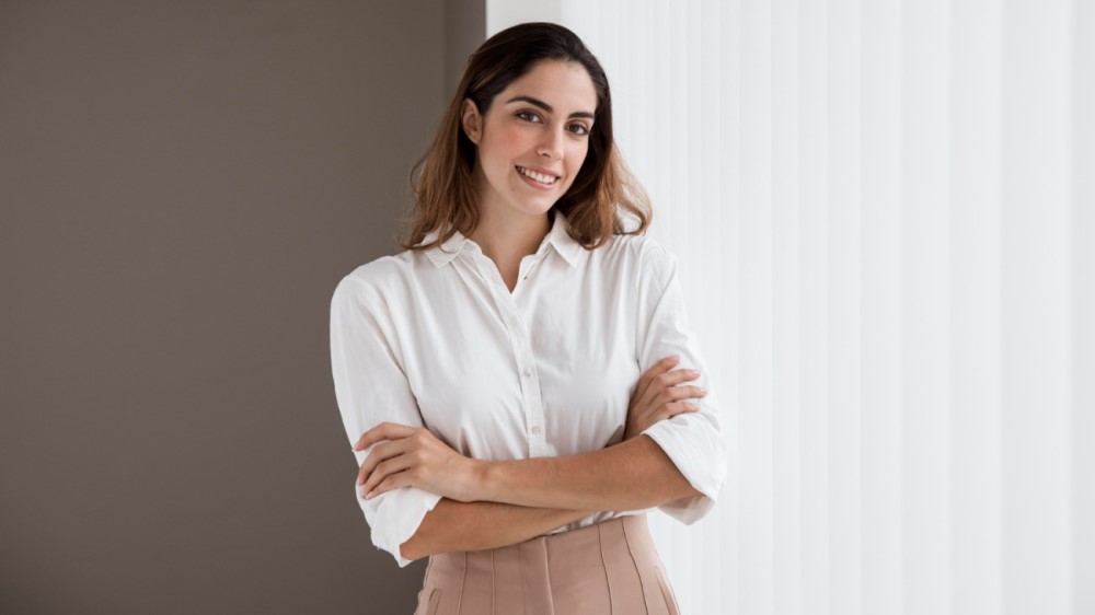 Uma mulher branca, vestindo camisa também branca, de braços cruzados e sorridente. Sua imagem transmite segurança, confiança e credibilidade, valores essenciais para qualquer profissional.