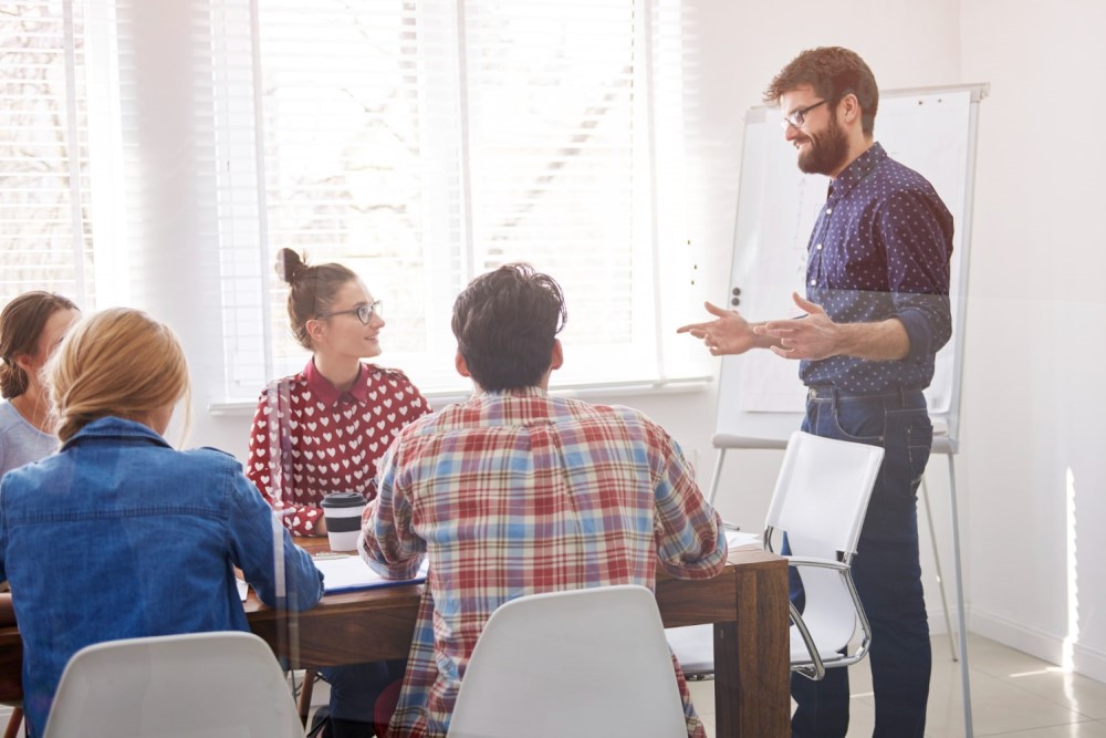 Um grupo de pessoas em uma sala de reuniões. A maioria delas está sentada ao redor de uma mesa e só um homem está de pé, falando com os demais.