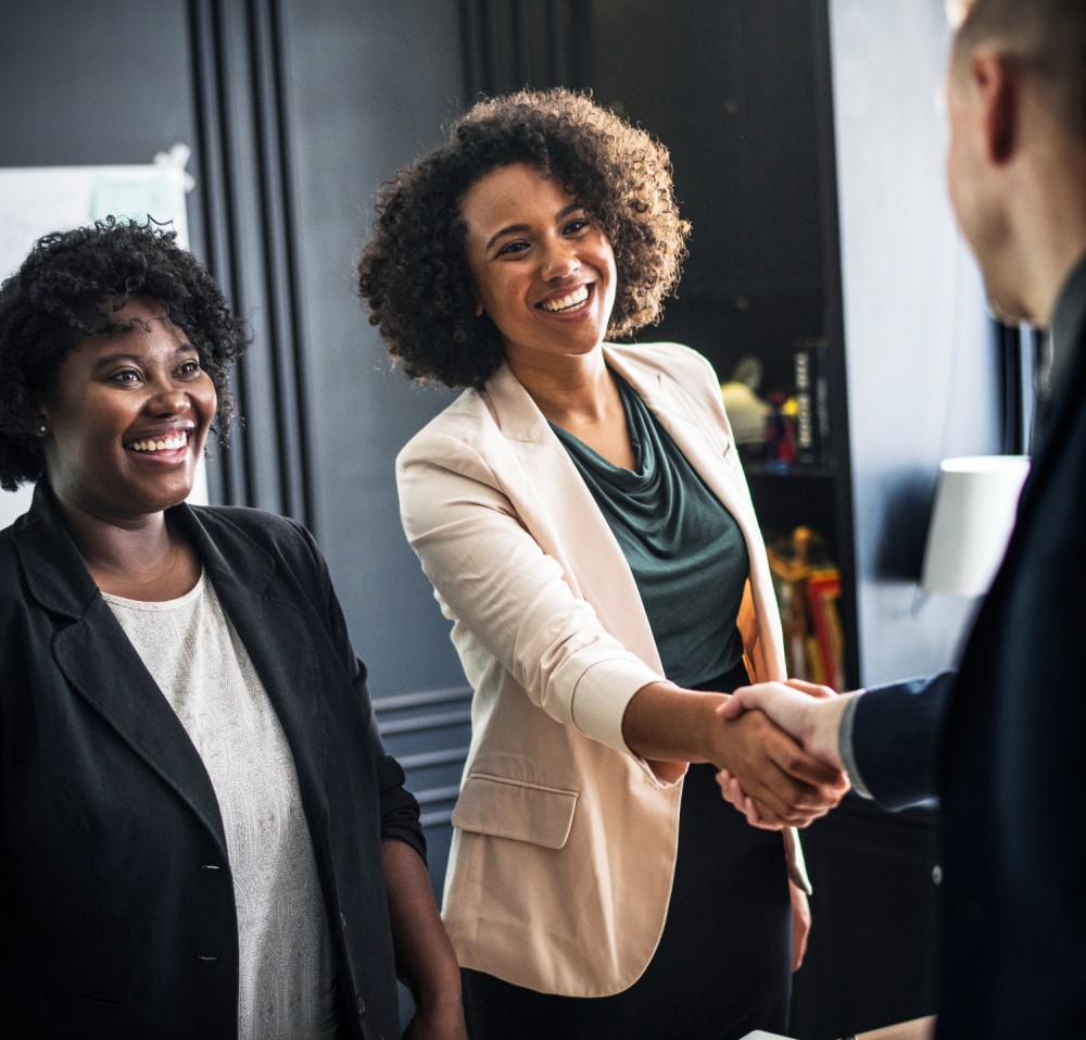 Duas mulheres negras, vestindo roupas sociais, sorriem. De frente para uma delas, no lado direito da imagem, há um homem de costas, um pouco desfocado, que estende uma das mãos a ela em sinal de “negócio fechado”.