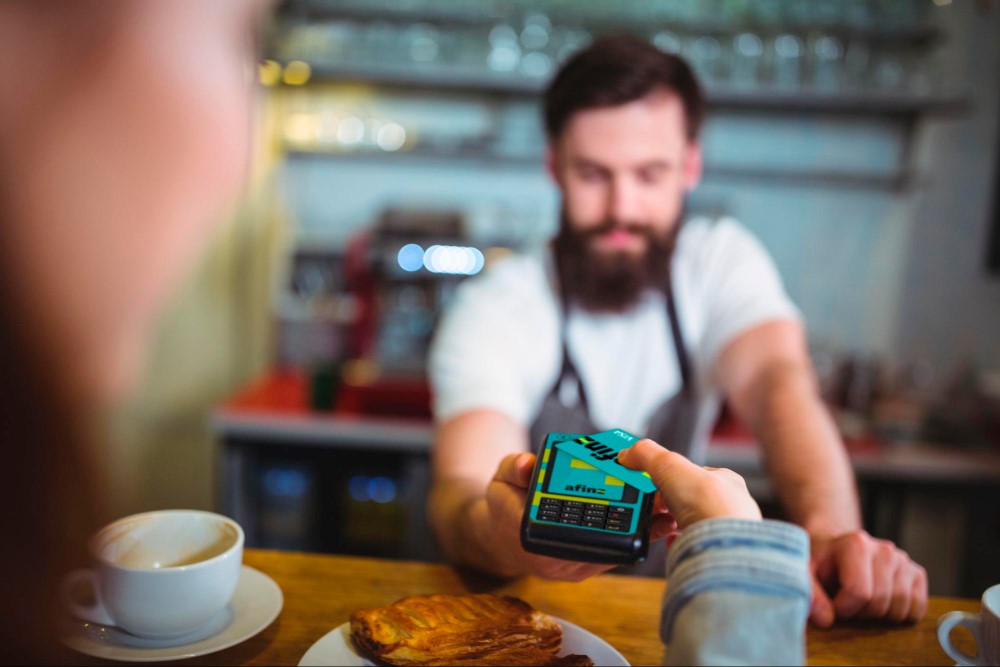 Mulher estendendo o braço para aproximar o cartão de crédito em sua mão, na maquininha. O comerciante a sua frente segue o mesmo movimento, segurando a máquina. Ao redor, vemos um pão e uma xícara de café, os itens que estão sendo comprados.