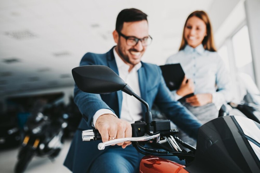Homem branco vestindo terno azul está sentado em uma moto e sorri enquanto segura o guidão com as mãos. Ao seu lado, vemos uma mulher sorridente representando uma vendedora.