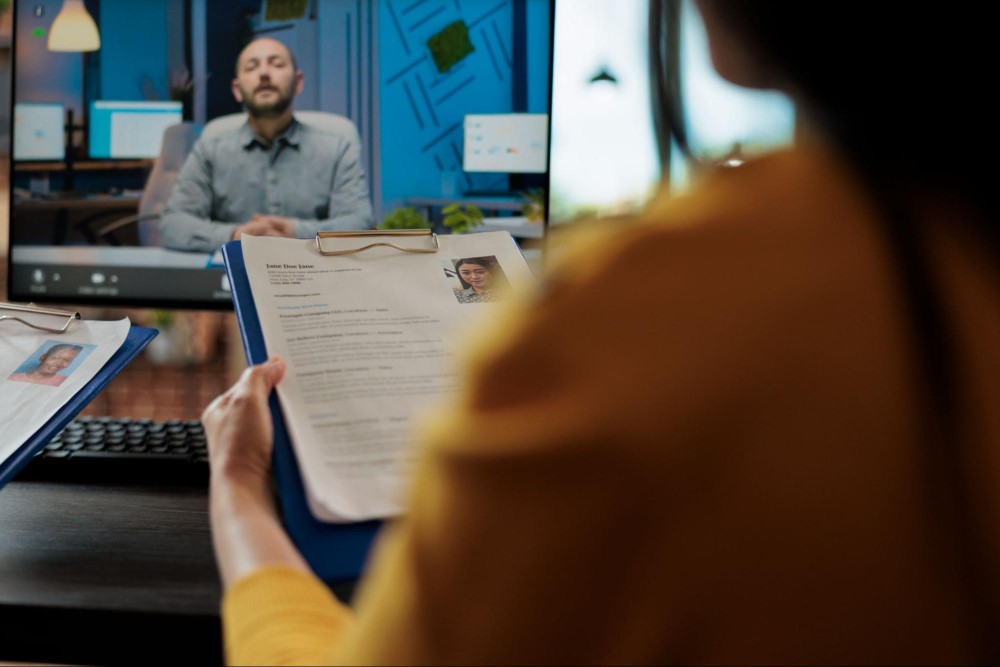 Mulher com camisa amarela está de costas para a imagem, enquanto segura uma caderneta com um currículo. À sua frente, vemos um monitor de computador onde está uma chamada de vídeo com um homem branco, vestido de social. Eles estão em uma entrevista de emprego online.