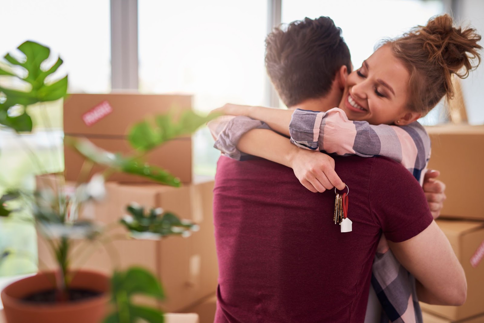 Na imagem um casal se abraça, a mulher sorri e segura a chave de uma casa. Ao fundo é possível ver uma planta e caixas de mudança.